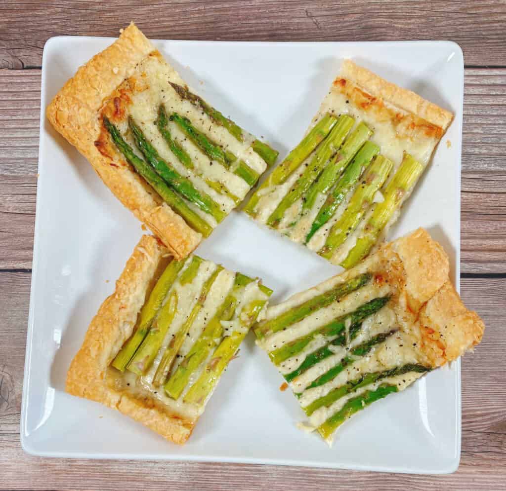 slices of asparagus and gruyere tart sit on a white square platter on a wooden background. There are 4 squares of the tart, two showing the tops of the asparagus and two showing the bottom of the asparagus spears. 