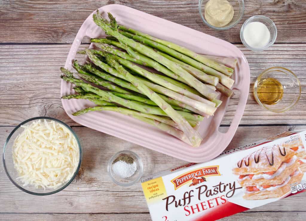 ingredients for asparagus and gruyere tart sit on a wooden background. Puff pastry, shredded gruyere cheese, salt and pepper, fresh asparagus spears, dijon mustard, honey and cream. 