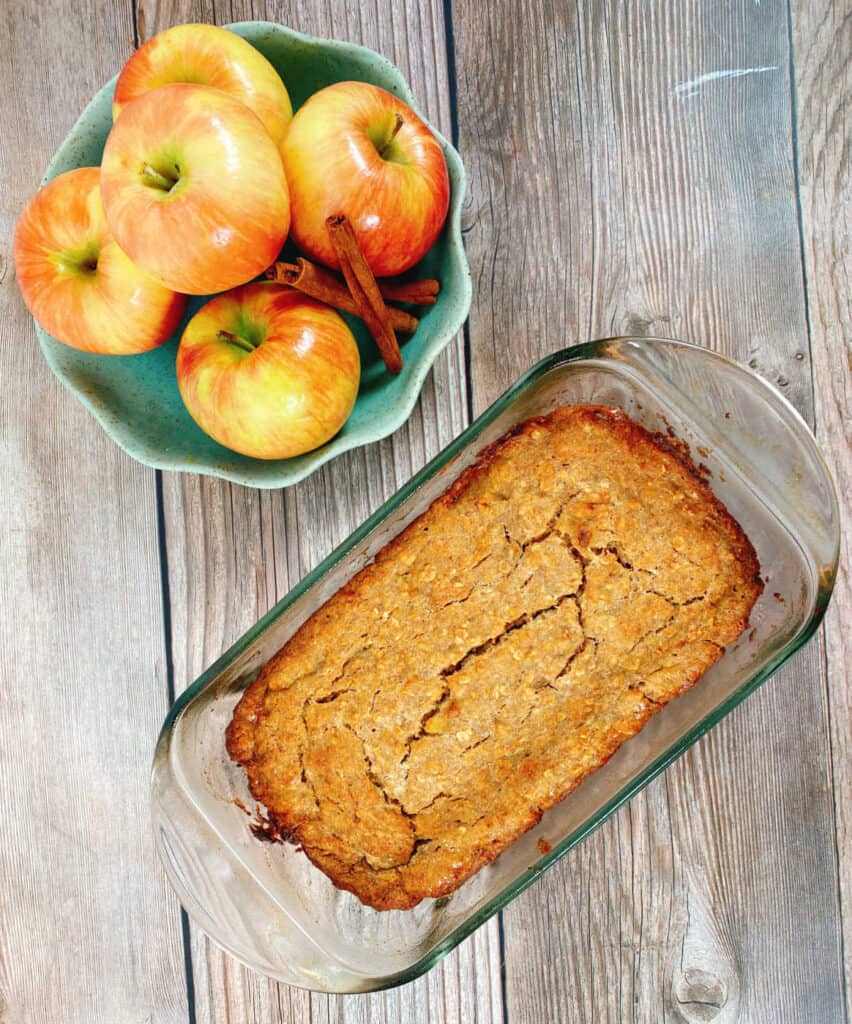 On a wooden background sits a bowl of honeycrisp apples and cinnamon sticks in a light green scalloped edge bowl. Below that sits the banana applesauce oatmeal bread, baked in a glass loaf pan. The top of the bread is cracked and golden brown. 