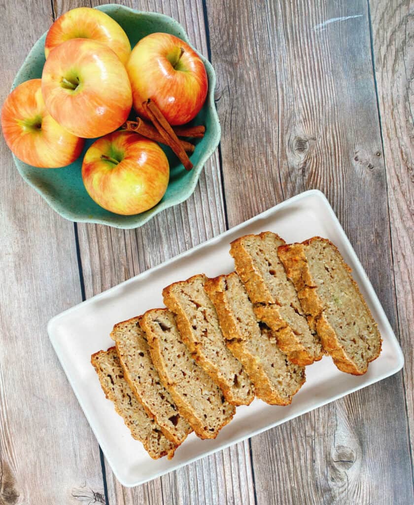 On a wooden background sits the banana applesauce oatmeal bread cut into slices on a rectangle white platter. There is also a bowl of honeycrisp apples and a few cinnamon sticks in a green bowl with scalloped edges. 