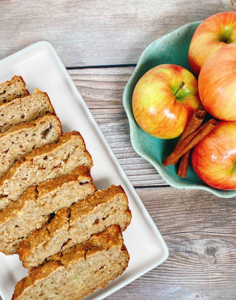 Close up image of the banana applesauce oatmeal bread. Slices of bread are layered on a white, rectangle shaped platter. Opposite the bread sits a light green bowl with scalloped edges. In the bowl are stacked honeycrisp apples and whole cinnamon sticks. 