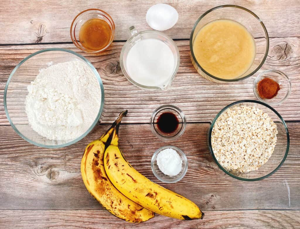 The ingredients for banana applesauce oatmeal bread sit on a wooden background. From left to right: all purpose flour, honey, bananas, almond milk, egg, vanilla, baking soda, applesauce, cinnamon and old fashioned oats. 