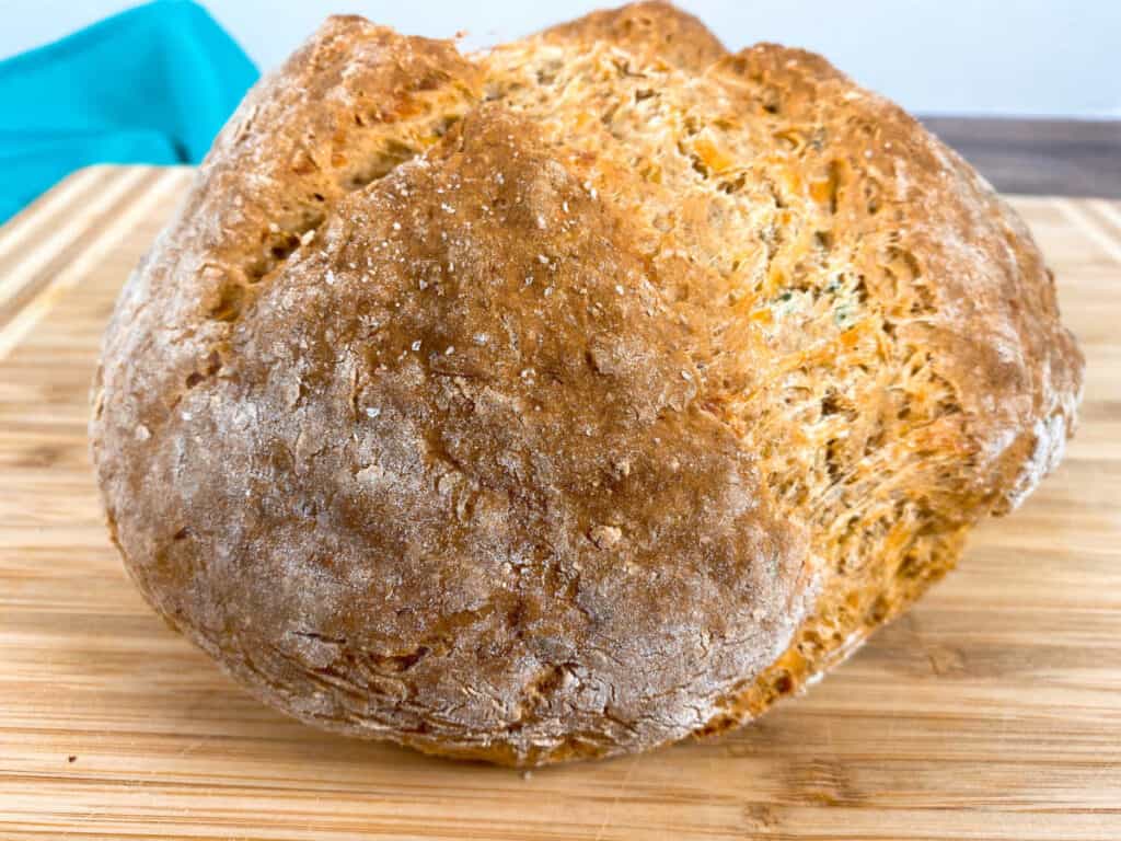 a freshly baked loaf of cheddar thyme irish soda bread sits on a wooden cutting board. the top of the bread is split, showing the baked goodness inside. 