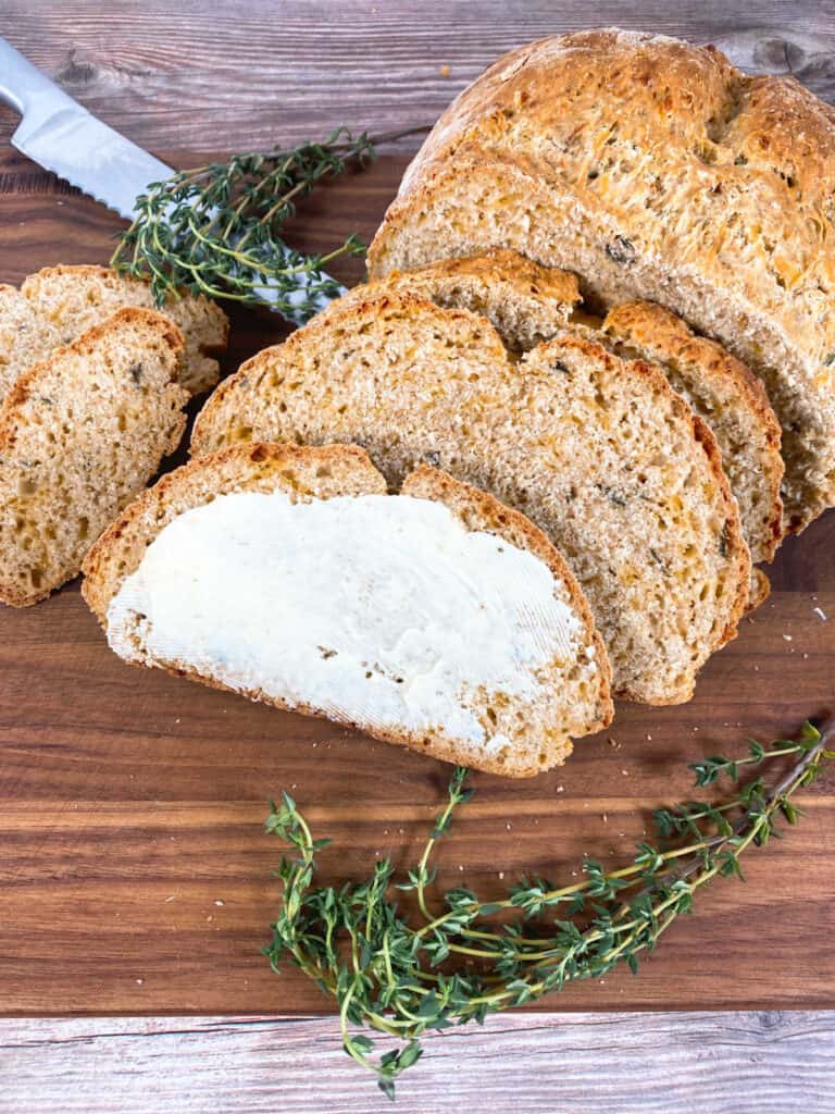 slices of cheddar thyme irish soda bread sit on a dark brown wooden cutting board. fresh thyme is arranged on the plate and a serrated knife sits in the background. The first slice of bread is buttered. 