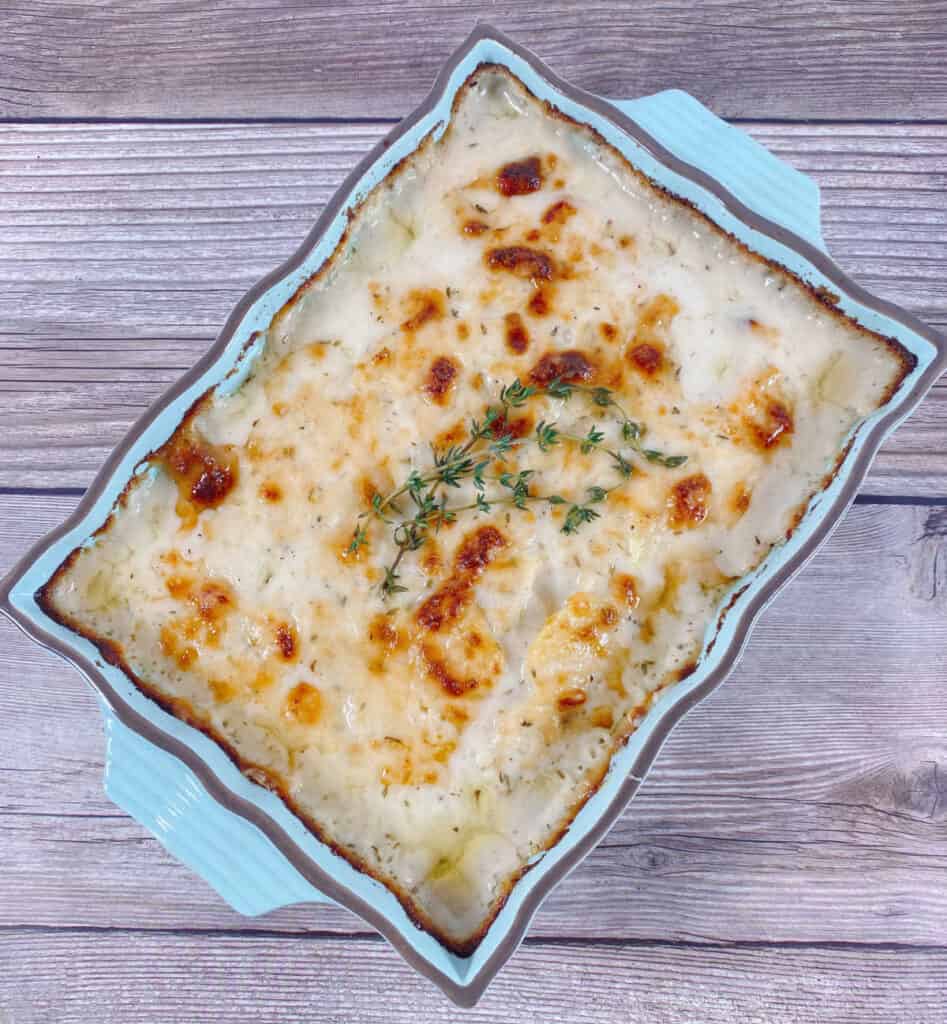a light blue casserole dish of scalloped potatoes sits at an angle on a wooden background. the scalloped potatoes are browned in spots from the crisping of the cheese and a few sprigs of fresh thyme sits in the center of the dish.