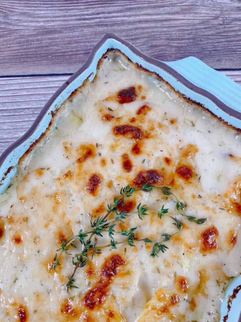 A close up image of a light blue casserole dish sitting at an angle on a wooden background. The dish is full of cheesy scalloped potatoes, browned in places from the cheese, and sprigs of fresh thyme sit on top. 