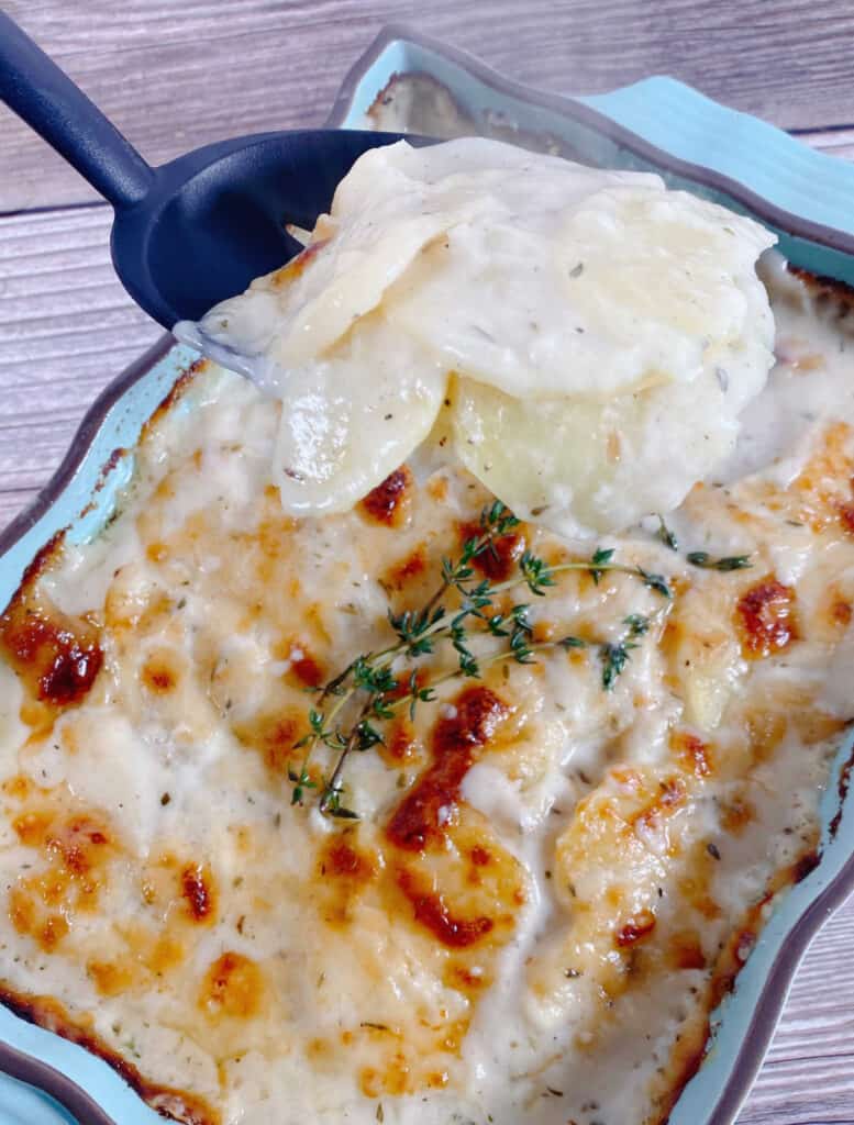 A slotted spoon lifts of a heaping spoonful of the easy cheesy scalloped potatoes from a light blue casserole dish. The dish sits on a wooden background. The remainder of the potatoes in the dish are lightly golden brown from the cheese and sprigs of fresh thyme sit on top. 