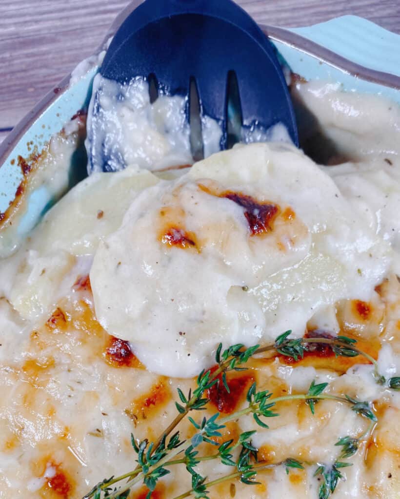 A slotted spoon lifts up a heaping spoonful of the easy cheesy scalloped potatoes from a light blue casserole dish. 
