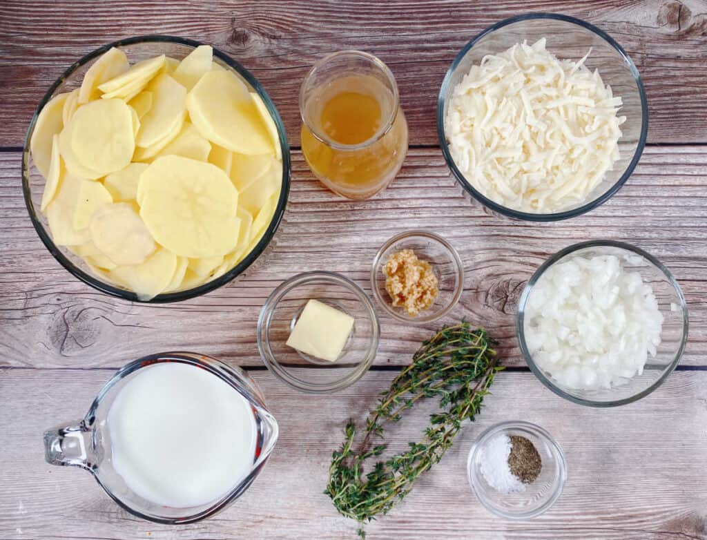Ingredients for Easy Cheesy Scalloped Potatoes sit on a wooden background. From left to right: a bowl of thinly sliced potatoes, a measuring cup of milk, a glass of chicken stock, a pat of butter, minced garlic, fresh thyme, a bowl of shredded cheese, a bowl of diced onion and a small dish of salt and pepper. 