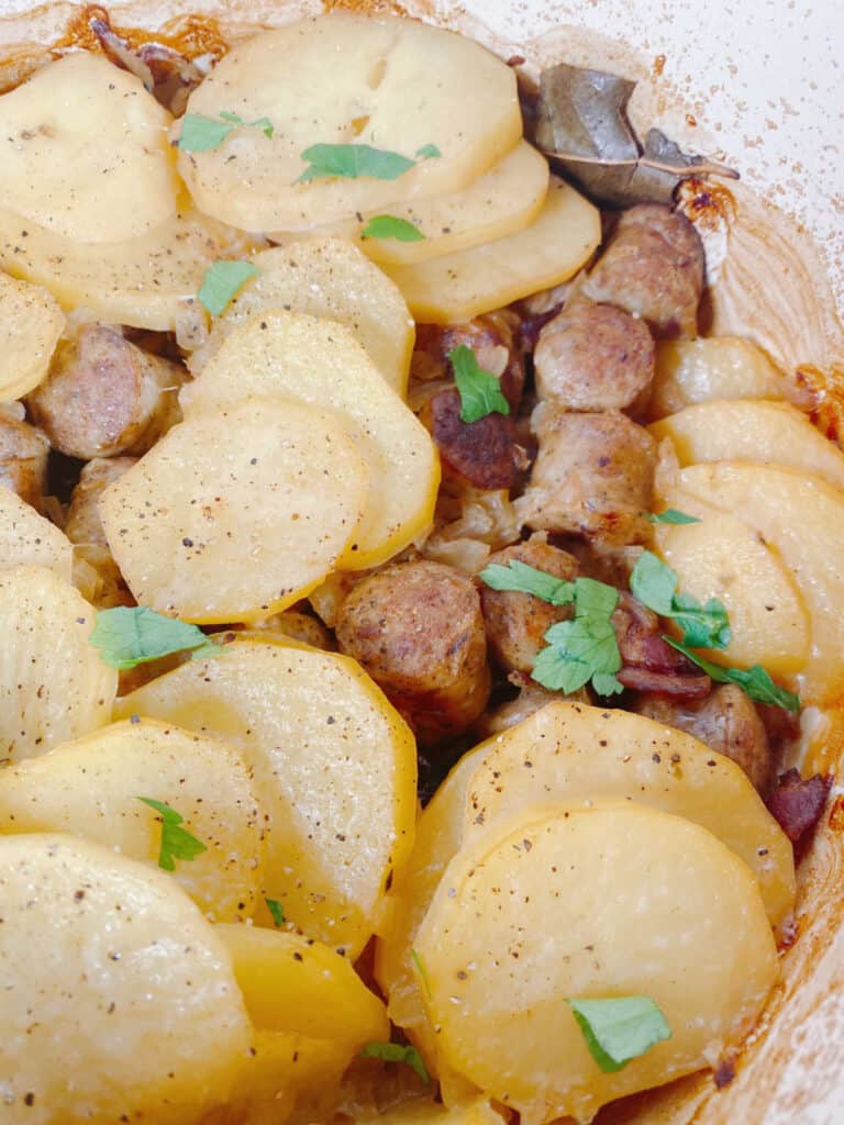 close up of easy irish dublin coddle in a dutch oven. Sliced potatoes, seasoned with salt and pepper and garnished with freshly chopped parsley cover most of the image. Pieces of browned sausage and bacon peek through the potatoes. 