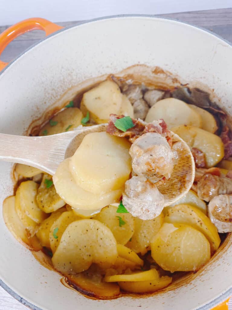 A wooden spoon lifts up the ingredients of this easy irish dublin coddle. On the spoon are cooked potatoes, sausage and bacon pieces. 