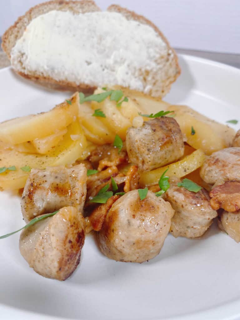 a shallow white bowl is full of easy irish dublin coddle - a dish made with sausage, bacon, potatoes and onions and garnished with fresh parsley. A slice of buttered soda bread sits in the background. 