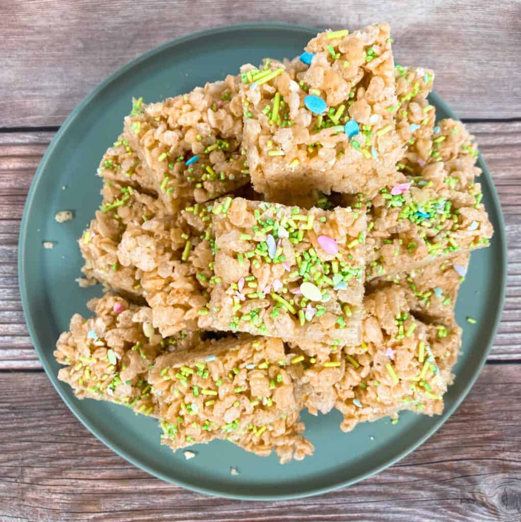 a stack of fruity easter rice krispie treats sits on a pale green plate on a wooden background. There're topping with easter sprinkles, in pastel colors. Some sprinkles are in the shape of an easter egg. 