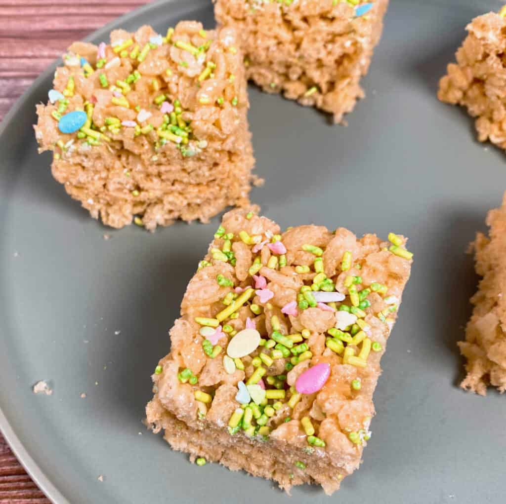 rice krispie treats made with fruit flavored marshmallows sit on a pale green plate and are topped with spring/easter sprinkles. 