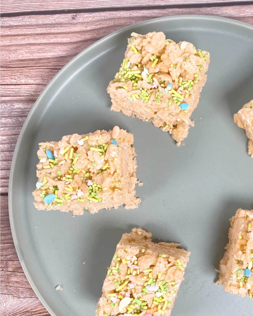 squares of fruity easter rice krispies sit on a pale green plate on a wooden background. the tops of the treats are covered in easter sprinkles, pastel in color and some are in the shape of easter eggs. 