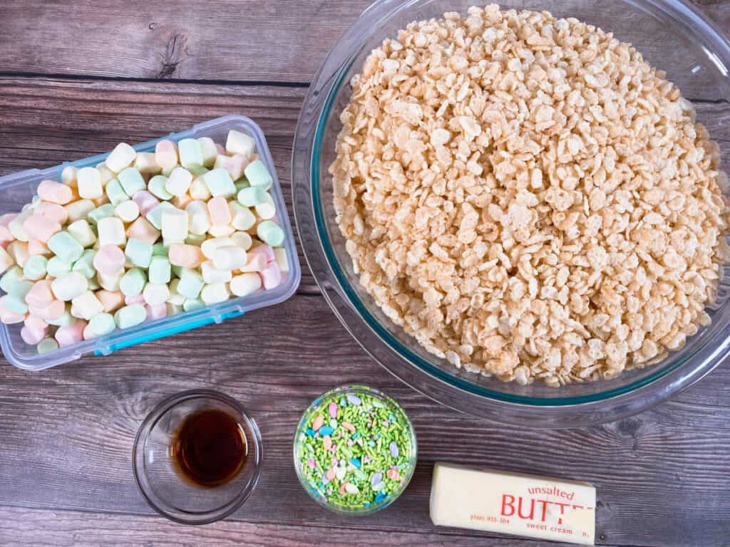 ingredients for fruity easter rice krispies sit on a wooden background. Fruity flavored marshmallows, rice krisipes cereal, vanilla extract, butte and easter sprinkles. 