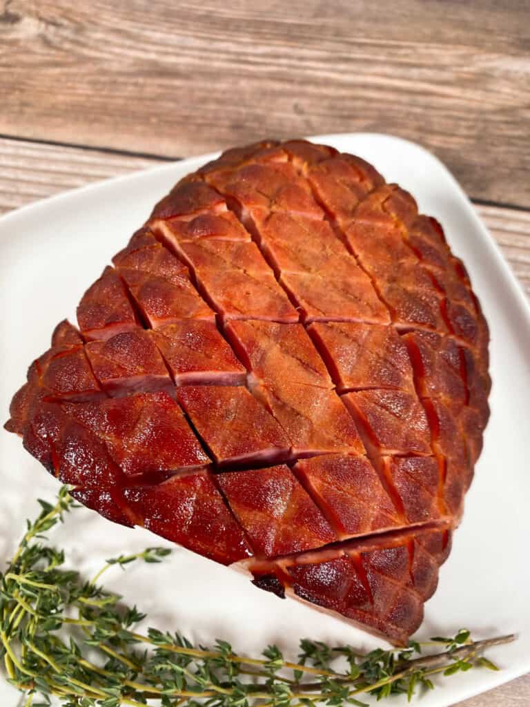 close up picture of honey bourbon baked ham sitting on a white plate garnished with thyme. the top of the ham is scored in a x pattern which allows the glaze to drizzle in. 