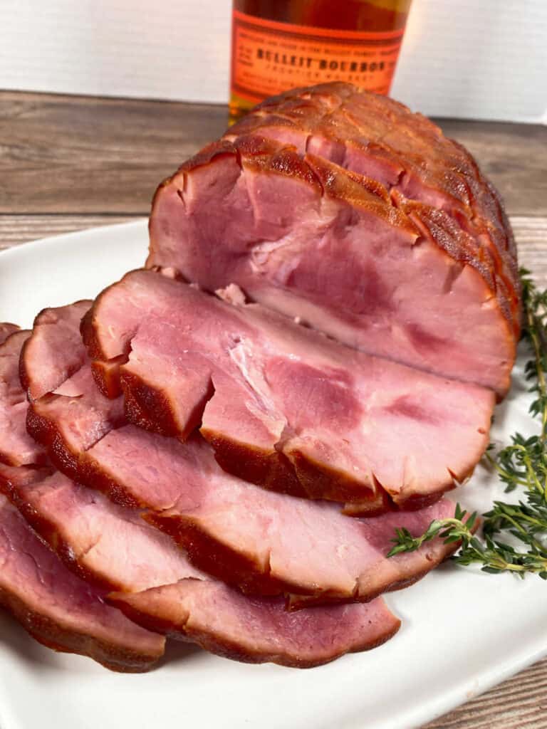 close up image of the slices from a honey bourbon baked ham sit on a white plate garnished with fresh thyme. 