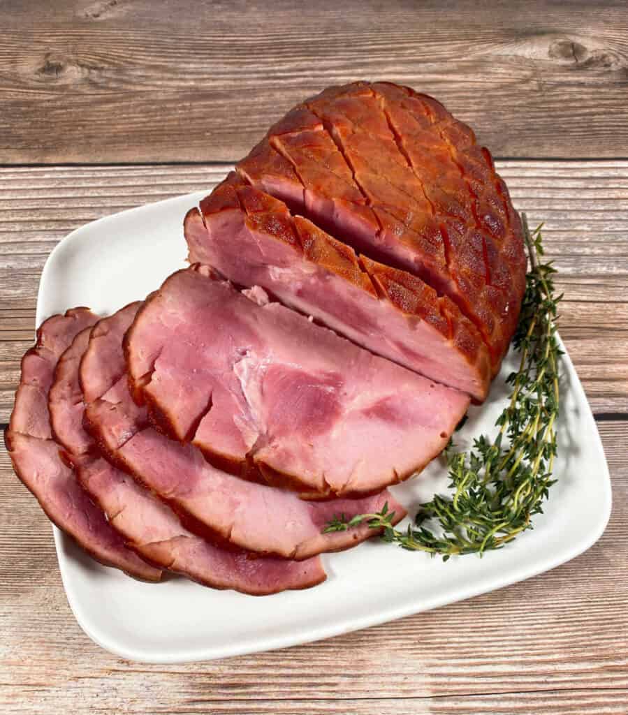 Slices of a honey bourbon baked ham sit on a white plate on a wooden background.