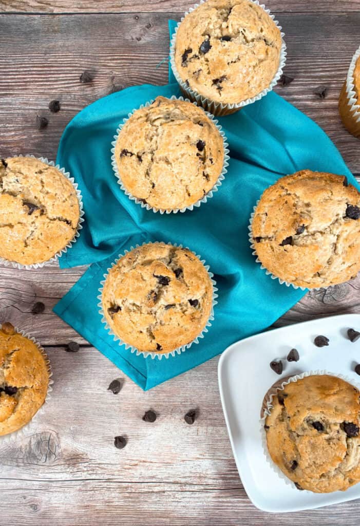 Image looking down. On a wooden background sit jumbo chocolate chip muffins. Some are arranged on top of a teal napkin, another on a white plate. Some are cut off from fully being in the image. Chocolate chips are scattered around the muffins. 