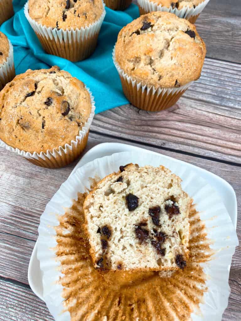 Image look down on a jumbo banana chocolate muffin, sliced in half. The muffin half sits on its wrapper and there are tons of chocolate chips studded in the muffin. Additional muffins sit in the background 