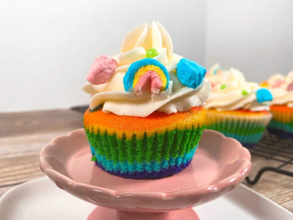 Rainbow vanilla cupcake sits on a pink cupcake stand with the other cupcakes behind it. Cupcake in the front is topped with vanilla buttercream frosting and is decorated with marshmallows from lucky charms cereal. 
