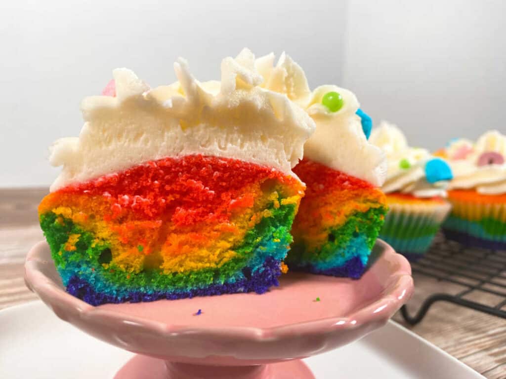Rainbow vanilla cupcake sits, cut in half to better show the colors, on a pink cupcake stand, The other cupcakes sit on a wire cooling rack in the background. 