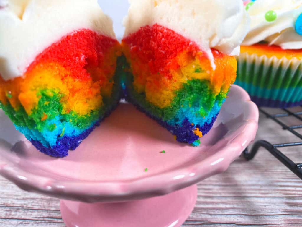 Close up image of the rainbow vanilla cupcake to show the color gradient in the cake. The cupcake is sitting on a pink cupcake stand and is sliced in half. 