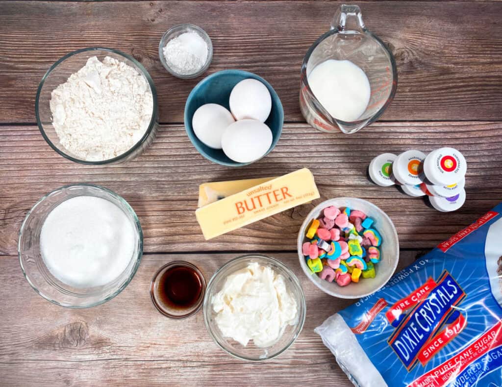 Ingredients for rainbow vanilla cupcakes sit on a wooden background. All purpose flour, sugar, butter, milk, sour cream, baking powder, vanilla and salt make up the cupcake ingredients. powdered sugar, food coloring and marshmallows from Lucky Charms cereal make up the frosting and decorations. 