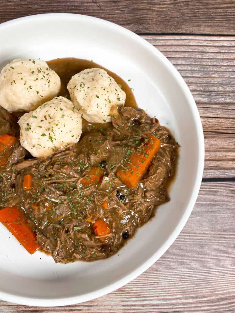 A shallow white bowl is full of sour beef and dumplings. The beef is marinated in vinegar and spices before being slow cooked with carrots and onions, then topped with  gravy made from gingersnap cookies. The potato dumplings are homemade and sprinkled with parsley. 