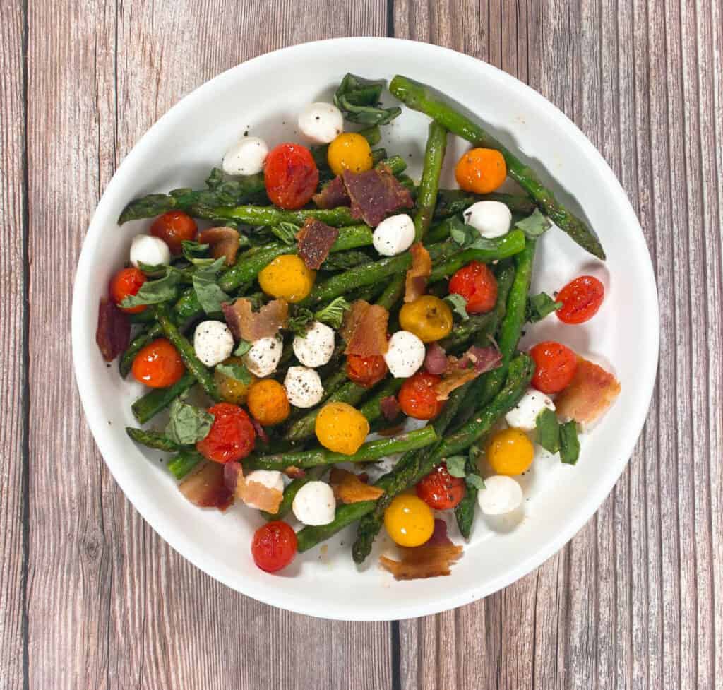 On a wooden background sits a white shallow plate full of the asparagus bacon caprese salad. The salad is made with asparagus, grape tomatoes, mozzarella, bacon, basil and balsamic glaze. 