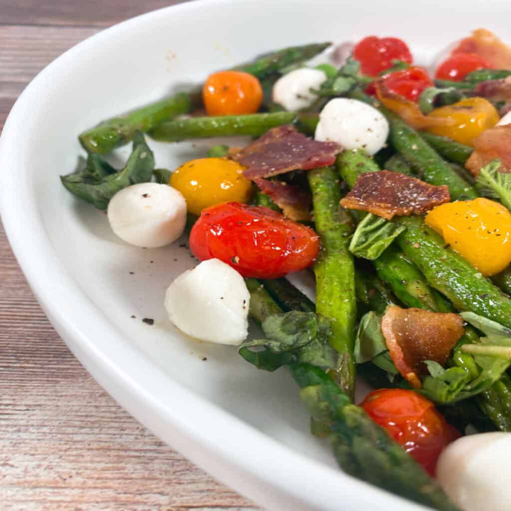 Close up side image of Asparagus Bacon Caprese Salad. Asparagus spears, grape tomatoes, crispy chopped bacon and mozzarella balls sit in the bowl, topped with chopped fresh basil and seasoned with salt and pepper. 