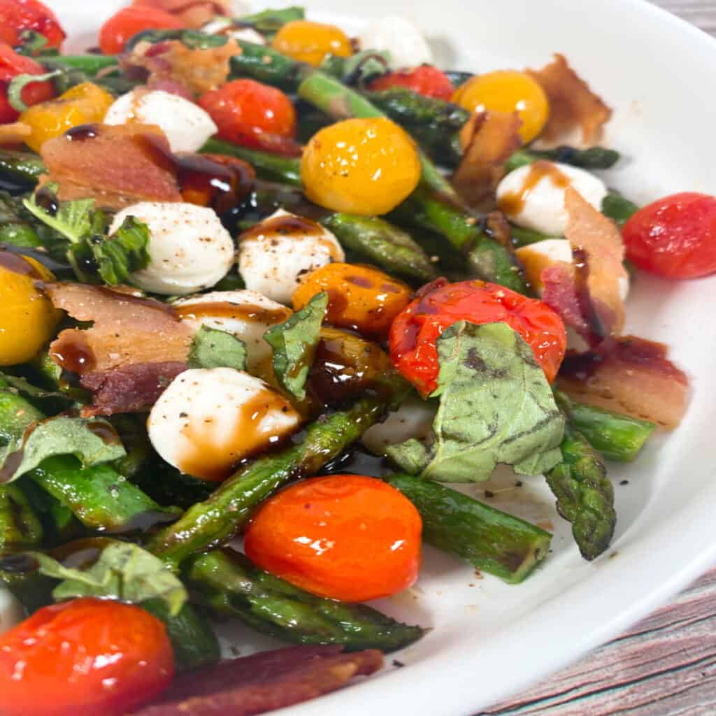 Close up image of Asparagus Bacon Caprese Salad. Spears of asparagus, grape tomatoes, mozzarella balls, chopped basil and crispy bacon are topped with balsamic glaze. The salad is in a white, shallow bowl. 