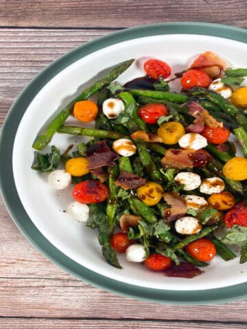 On a wooden background sits a white shallow bowl on top of a green plate. The bowl is filled with asparagus bacon caprese salad, made with asparagus, grape tomatoes, mozzarella balls, fresh basil and crispy bacon. It's seasoned with salt and pepper and drizzled with balsamic glaze.