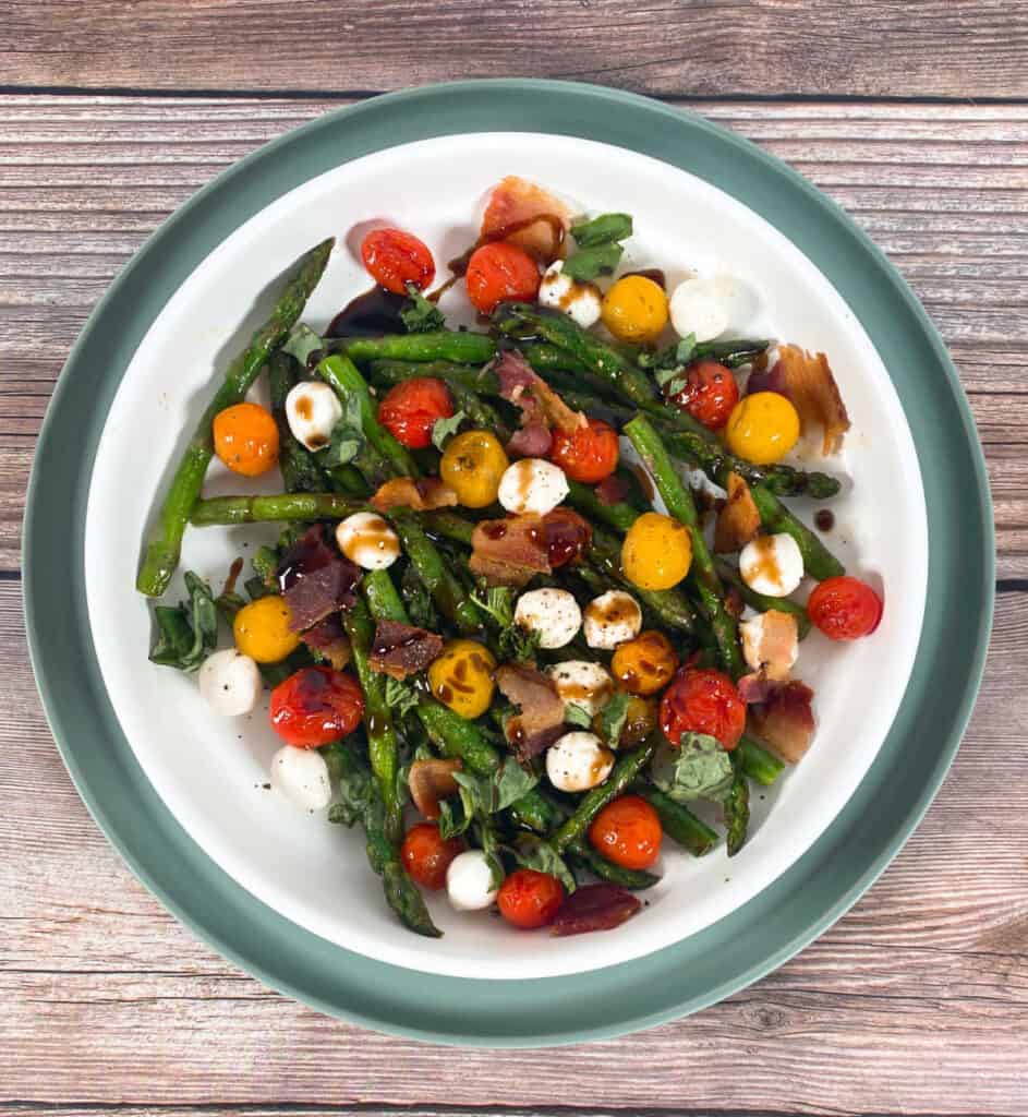 Image is top view looking down. On a wooden background there is a light green plate with a shallow white bowl on top. The Asparagus Bacon Caprese salad is in the white bowl, drizzled with balsamic glaze.