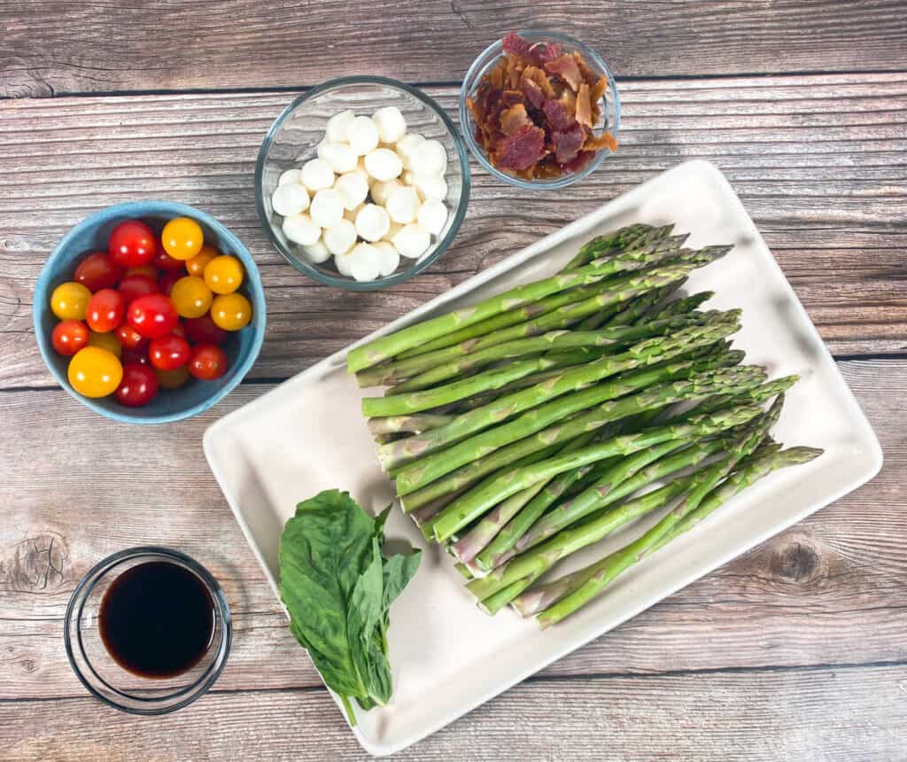 Ingredients for asparagus bacon caprese salad sit on a wooden background. Ingredients include balsamic glaze, grape tomatoes, mozzarella balls, chopped bacon, asparagus and fresh basil. 