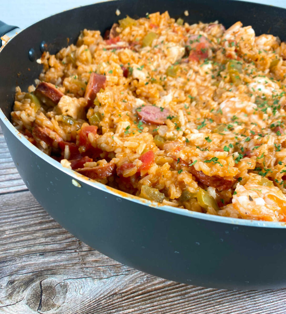 Side view of a large pot of jambalaya sitting on a wooden background. The top of the dish is sprinkled with parsley and the shrimp, chicken and sausage are pushed to the top. 