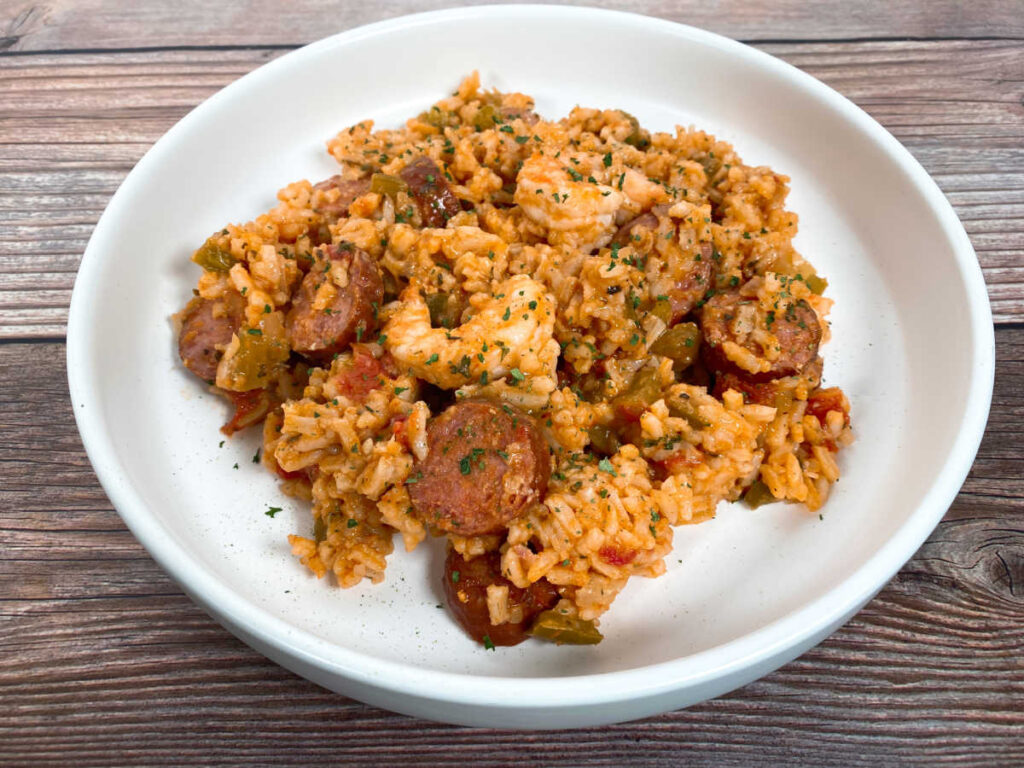 Top down image of the jambalaya, sitting in a shallow white bowl on a wooden background. The dish is sprinkled with parsley. 