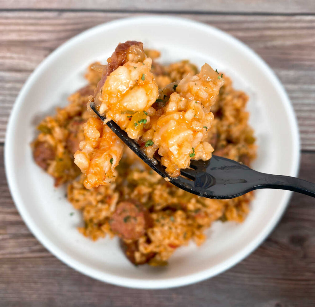 A black fork with shrimp, rice and sausage is held above a bowl of the jambalaya. The bowl below is shallow and white and filled with the remaining dish. It is slightly out of focus. 