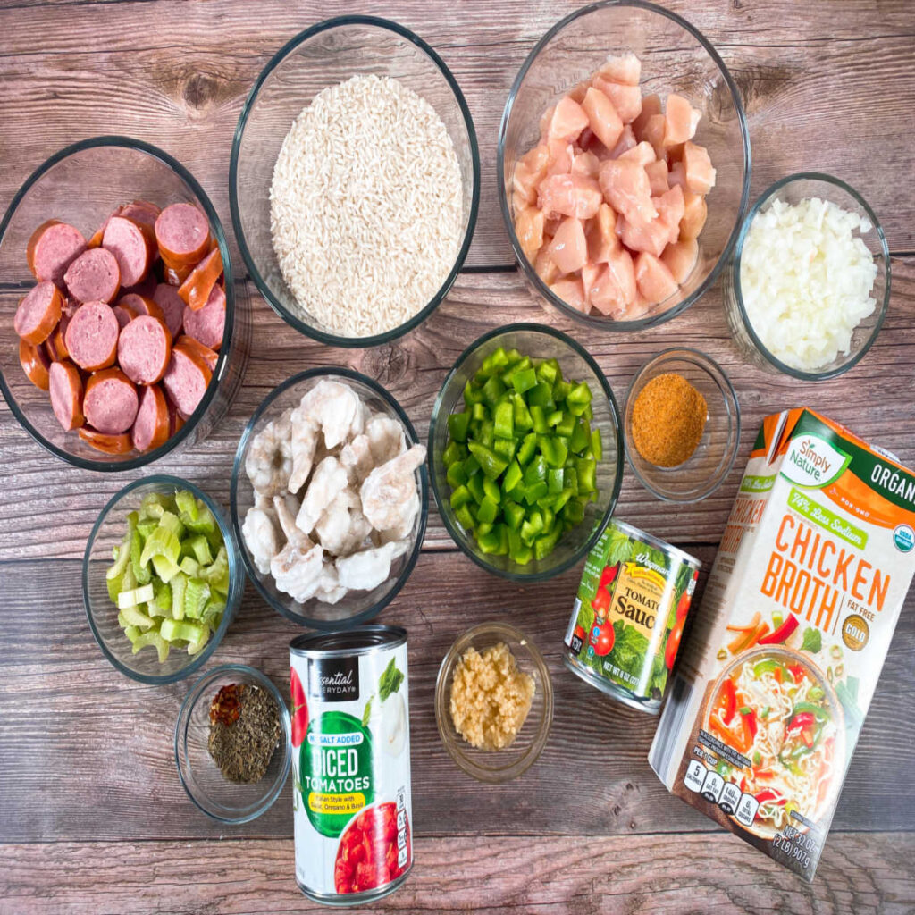 Ingredients for this recipe sit on a wooden background. Ingredients include bite sized pieces of chicken, sausage sliced into rounds, shrimp, green pepper, celery, onion, minced garlic, canned diced tomatoes, tomato sauce, chicken broth, Cajun seasoning, long grain white rice, dried thyme and oregano, crushed red pepper flakes, worcestershire sauce, salt and pepper. 