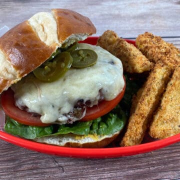 close up of burger in a red basket. Top pretzel roll is tilted off the burger to show the toppings and cheese on the burger.