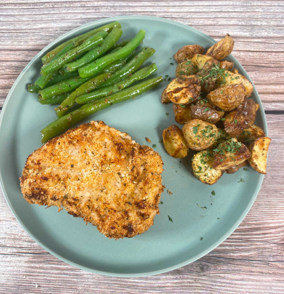 overhead image of the crusted pork chop on a round, green plate. Potatoes and green beans are on the plate as well. 