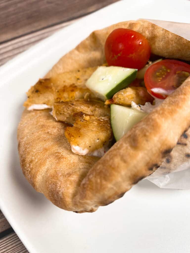 close up side image of the gryo - chicken sits on a pita spread with tzatziki sauce, topped with chopped cucumbers and tomatoes. 