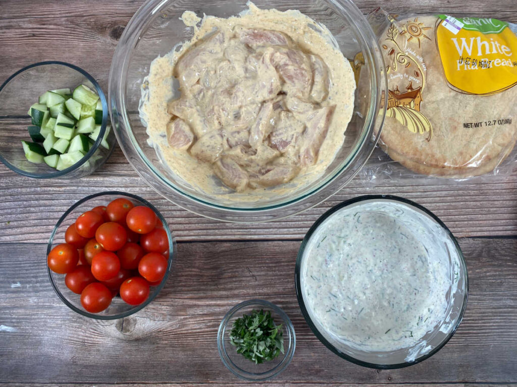Ingredients sit in glass bowls - prepared tzatziki sauce, marinating chicken, grape tomatoes, chopped cucumbers, parsley and pita bread. 