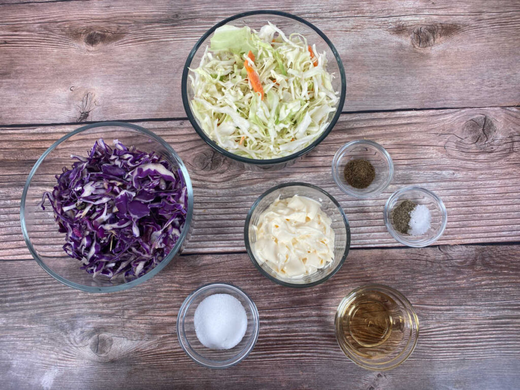 ingredients for this recipe sit in glass bowls on a wooden background. Ingredients include coleslaw mix, shredded red cabbage, mayo, sugar, apple cider vinegar, celery seed, salt and pepper.