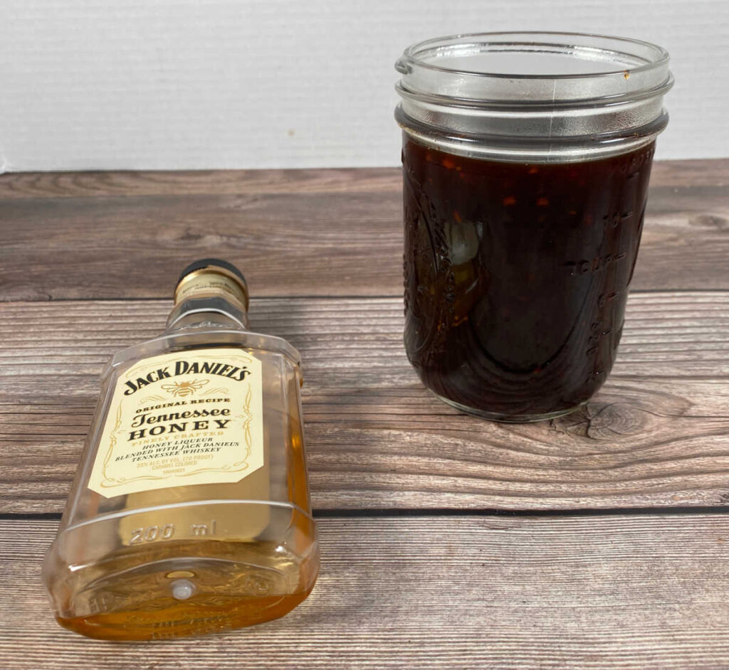 bbq sauce sits in a mason jar with a bottle of jack daniels next to it on a wooden background. 