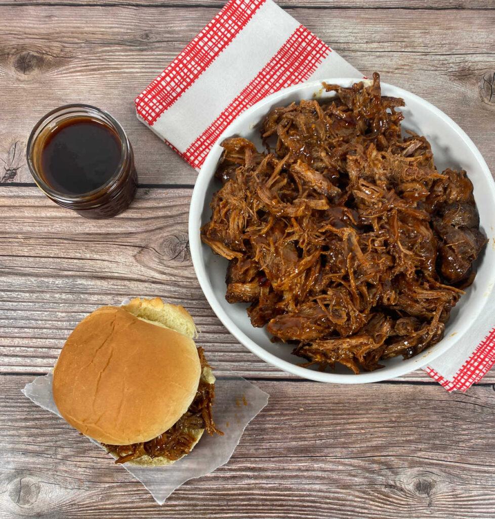 overhead view of a pulled pork sandwich sitting on a wooden background. Plate of additional pork sits in a white bowl on a checkered napkin and a mason jar of the bbq sauce sits in the background. 