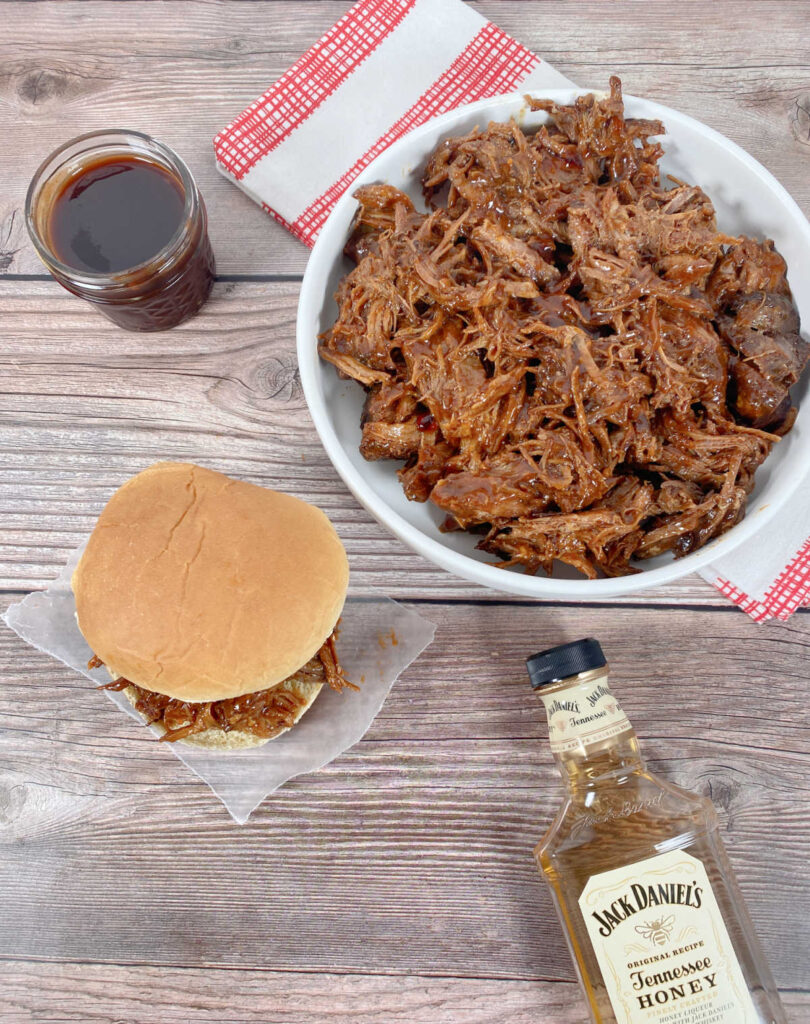 Overhead view of the pulled pork sandwich sitting on a square of wax paper. Additional pulled pork sits in a bowl, next to it the homemade bbq sauce and below it, the bottle of liquor used in the sauce. 