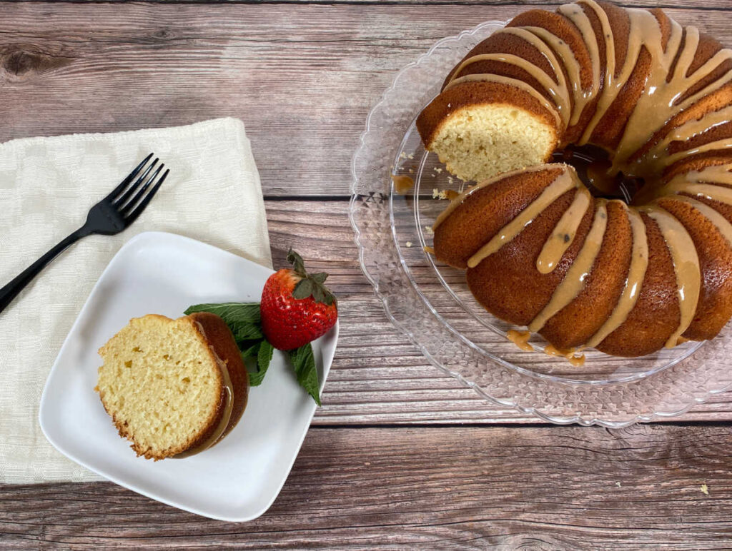 Slice of hot milk cake sits on a square white plate with a strawberry, napkin and fork. On the right is the remainder of the cake on a glass dish. 