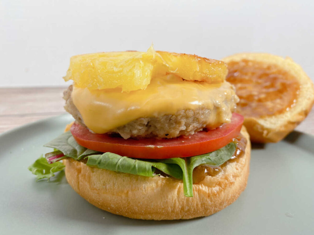 burger sitting on a light green plate. Top of burger bun, covered with teriyaki sauce is in the background. 